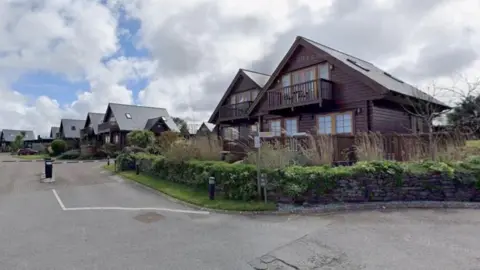 The image shows several wooden holiday lodges with pitched roofs. There are seats on the verandas and a balconies on the first floor of the lodges, which are surrounded by shrubs.
