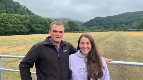 Ioan Jones and Sara Jenkins, winners of Channel 4's Our Dream Farm - standing in front of a metal gate, with their farm - Llyndy Isaf - in the background.