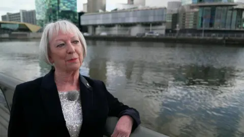 June Batty with bobbed white hair wearing a silver sequin top underneath a black velvet blazer. She has her arm resting on railing close to the water at Salford Quays with the Lowry in the background. She is smiling.