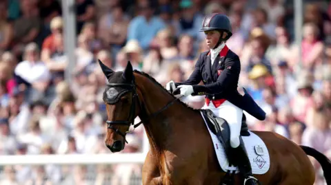 PA Media Ros Canter, dressed in blue and white equestrian clothes and a dark blue helmet, rides a brown horse in front of a crowd at the Badminton Horse Trials 2024.