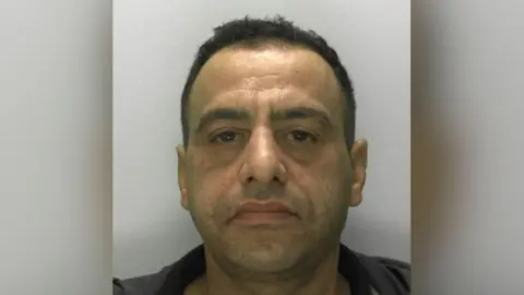 A police custody photo of James Brown. He has short dark hair, thick dark eyebrows and is wearing a dark top and hooded jacket.