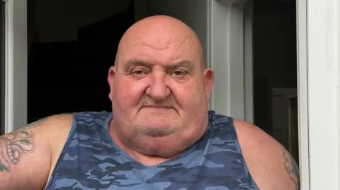 Graham Horn, who is wearing a blue coloured camouflaged vest and with tattoos on both arms, stands in his doorway. 