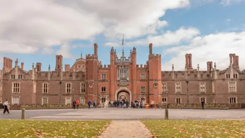 Historic Royal Palaces The front of a Tudor castle with an arched gateway entrance and a flag on top. Visitors and tourists are walking around a large courtyard at the front