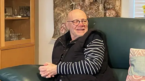 Malcolm wears a stripy navy and white top under a black gilet and sits on a green sofa. He is smiling and wears glasses.