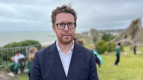 George Carden/BBC Man in blue jacket and white shirt with glasses, beard and ginger hair on a cliff top