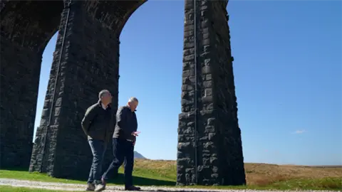 Nick Wooley/BBC Correspondent Danny Savage and Peter Myers walk below the viaduct, in the background are green rolling fields and a clear blue sky