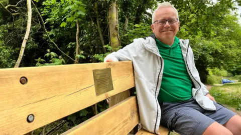 An older man with white hair sits on a wooden bench, which has a gold coloured memorial plaque on it, in a woodland area