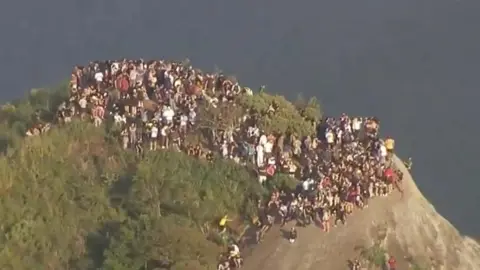 TV Globo Aerial photograph shows a large group of people standing at the top of a high point in Rio de Janeiro, Brazil. It's early morning, and the shadows are long, indicating sunrise.
