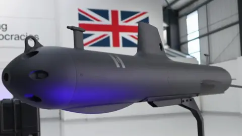 The image shows an unmanned submarine glider displayed indoors on a stand. The glider has a sleek, torpedo-like design with a matte dark finish and a pointed nose. It includes a vertical stabiliser fin and a long tail section for hydrodynamic control. A soft blue light is illuminating part of the body. In the background, there is a large Union Jack flag mounted on the wall and the setting appears to be a modern industrial or research space with white walls and high ceilings.