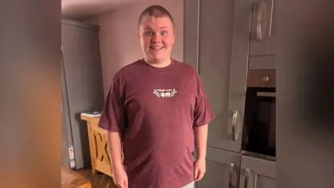 Leanne McCrotter A tall teenage boy wearing purple t shirt stands and smiles at the camera. he has very short brown hair, blue eyes and in the background is a family kitchen.