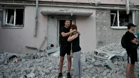 Couple stands at the foot of building strewn with debris