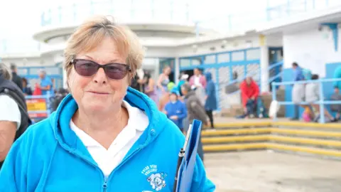 Sally Minty-Gravett wearing a blue hoody and holding a clipboard. She has short brown hair and glasses. 