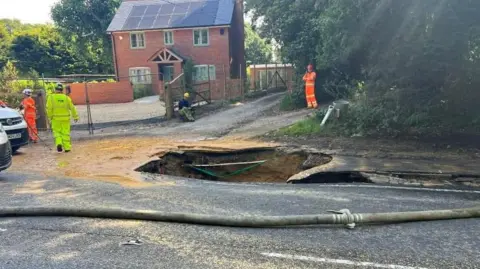A large hole in the road with three workers in hi-vis clothing working nearby, and a hosepipe sitting alongside the hole on the roadside