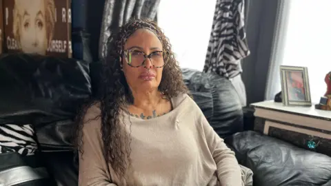 Lisa-Lynne Beckman is wearing a beige top, glasses and a headband sitting on a black leather sofa in front of her lounge windows