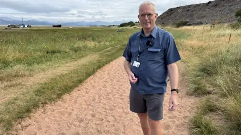 A middle aged man with short grey hair, wearing spectacles, a blue short-sleeved shirt, and grey shorts. He is standing on a sandy footpath cutting through rough scrabbly grass. Behind him, and to the left can be seen a tidal area with some boats which appear to be grounded, and mountains stretching into the distance. To the right is a high bluff.
