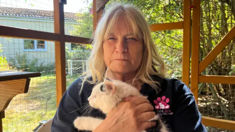 A woman with blond shoulder-length hair frowns as she holds a white cat inside a large wood and wire pen, with a green garden and house in the background. She is wearing a blue shirt with pink "Lincs Ark" logo in the shape of a paw.