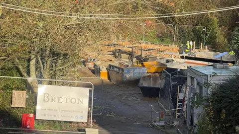 A construction site. There are temporary metal fences up. There are skip bins and a yellow construction vehicle. There is a person in green hi-vis working. There are green trees and bushes. 