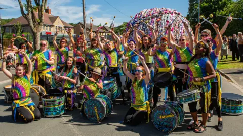 Bradford Council A group of women of different backgrounds pose with African drums. They are all dressed in colourful, vibrant outfits in blue, pink, yellow and green. They hold their arms in the air and in their hands are drumsticks which they pose with. Behind them is a giant ball of yarn, and groups of people are standing around. It looks like they are in a residential street and are part of a community event. 