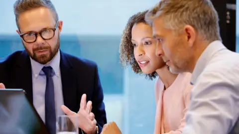 Getty Images A man in a suit and tie showing a man and a woman in smart clothing a laptop screen in an office environment 