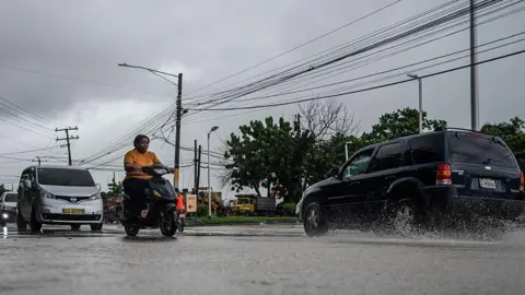 AFP via Getty Images Cars and a motorbike drive on a street in the Dominican Republic amid rain. Power lines can be seen overhead 