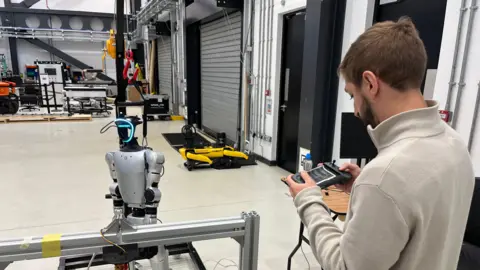 A robotic engineer is working with a prototype of a humanoid robot in a large laboratory at Heriot Watt University