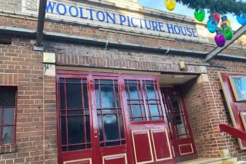 Art deco front of Woolton Picture House, which has its name sign in blue at the top of the single-stroey red brick building and red doors. Multi-coloured light bulbs hang from a green Christmas garland.