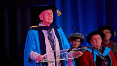 University of Huddersfield Former Huddersfield MP Barry Sheerman stood at a lectern. He is wearing a suit and tie with a blue cape over and a black cap with a yellow tassel. At the side of him are three people sat down who are wearing black caps.