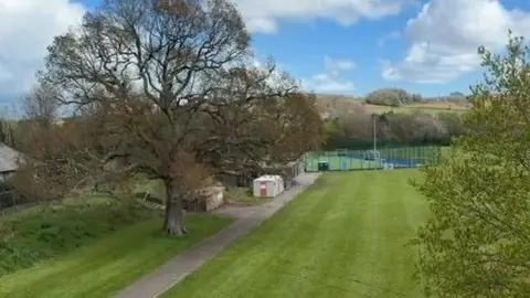 A large playing field with several buildings towards with back, with tennis courts. A large tree is in the field, with another to the side. 