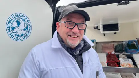 Nick looking into the camera and smiling, he's wearing a white coat over the top of his winter jacket. He's stood in front of his van, and the back doors are open so trays of fresh fish and seafood are visible. Over his right shoulder is a sign that reads 'The National Federation of Fishmongers 2022.'