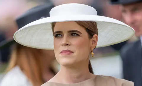 Princess Eugenie wearing a large wide-brimmed white hat, gold toadstool earring and brown dress
