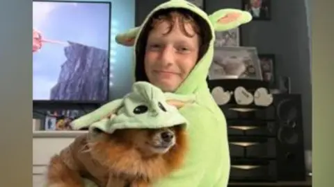 A boy in a green Shrek hoodie holding a dog which is also in a green shrek outfit - they are in a living room of a house with a TV in the background and the boy is smiling.