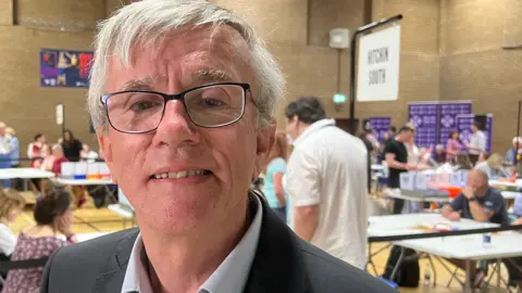 Steve Jarvis with short white hair smiling at the camera while wearing a grey jacket and pale blue shirt. There are tables behind him with people sitting behind them at an election count in a sports hall.