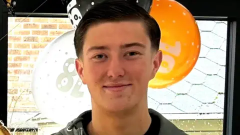 A young man with short brown hair smiles to camera. In the background celebratory balloons can be seen with 18 written on them.