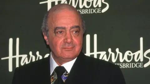 BBC Mohamed Al Fayed in a suit stood in front of a sign that says Harrods.