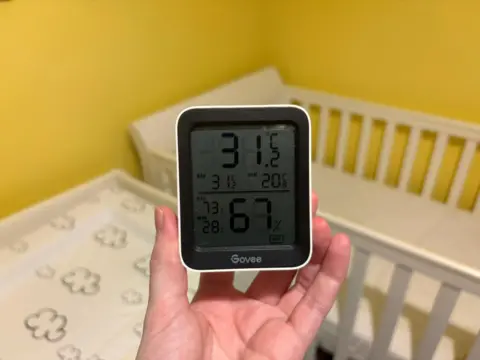 Jackson Bylett A digital thermometer is being held up in front of a crib. It shows a temperature of 31.2C