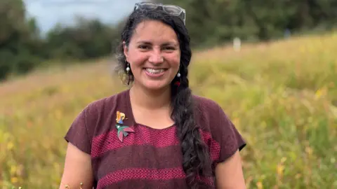 Malú is smiling and looking into the camera. She's standing in a filed of flax. She has her long black hair tied in a braid. Glasses are perched on top of her head. She's wearing a bird brooch and another brooch on her top.