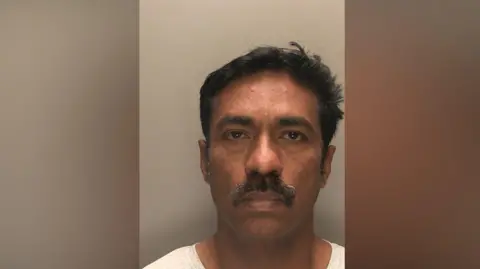 A police head and shoulders photo of a man with short black hair and a moustache, pulling a blank expression and wearing a white top against a grey wall.