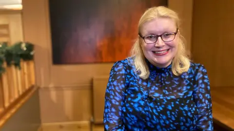 BBC Laura with blonde hair and glasses smiling as she looks into the camera. She has a blue and black patterned top on and is sitting in the corner of a room with a large picture on a wall behind her.