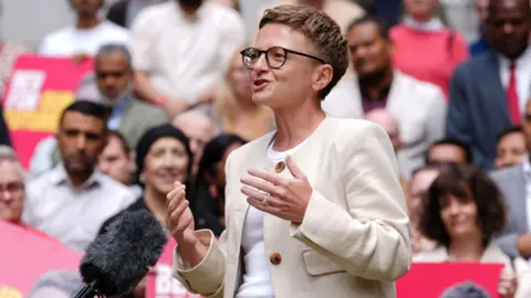 A woman with short dark hair giving a speech, she has glasses and is wearing a cream blazer and white top