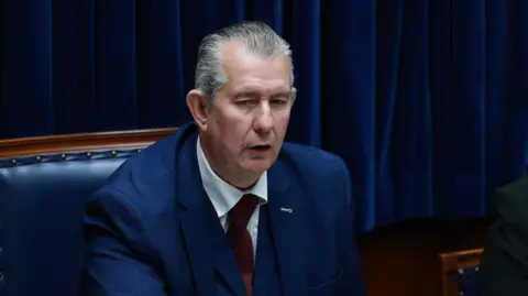 Edwin Poots has short grey hair and is wearing a blue blazer, red tie and white shirt.