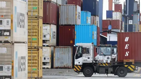 A white truck in front of stacks of different-coloured cargo containers at a port