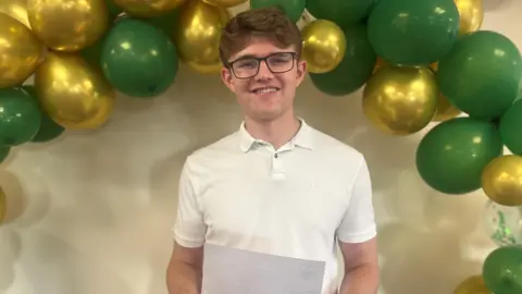 BBC Leo Crowe wears a white shirt and dark spectacles. He holds a piece of paper with his results and stands under an arch with green and gold balloons.