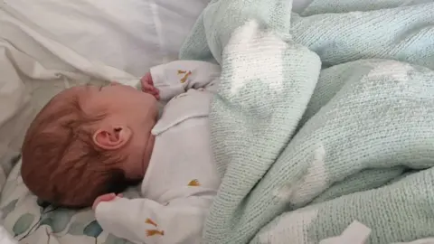 Joel Priddey Dark-haired baby lying in a Moses basket, wearing a white baby grow and teal blanket over her.