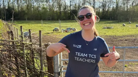 Sophie Martin standing in front of a gate to a field with sheep grazing. She is a young woman, with dark hair tied back, wearing sunglasses and smiling.