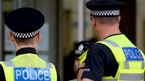 Image of the back of two police officers. They're wearing black police tops and florescent vests and police hats. 