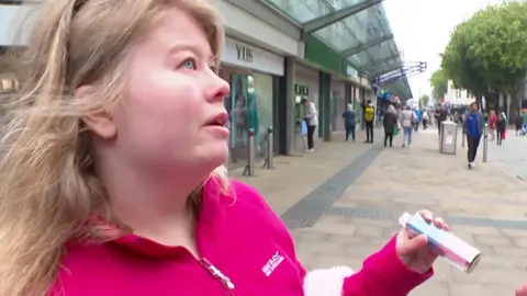 Blonde woman wearing pink fleece holding a pink and blue disposable vape.