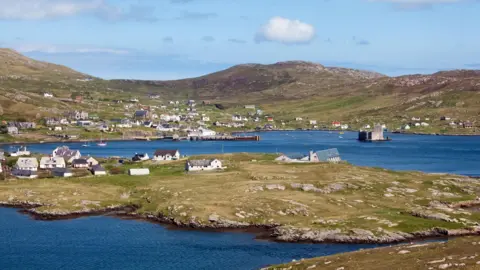 A view across Castlebay in Barra. A rugged coastal landscape is dotted with white-walled houses. In a sea loch is Kisimul Castle.