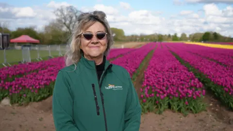Shaun Whitmore/BBC Niki Ellis has fair wavy hair and is wearing a green jacket, branded with Tapping House. She is smiling and standing in front of rows of tulips.