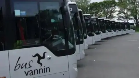 A row of silver Bus Vannin single-decker in a semi-circle. The have dark tinted windows and the b\us Vannin logo in black on the side.