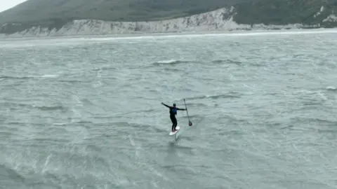 Emma Bridge A lone rider balances on a hydrofoil board while holding a paddle, skimming above choppy grey water with white cliffs stretching along the coastline in the background.